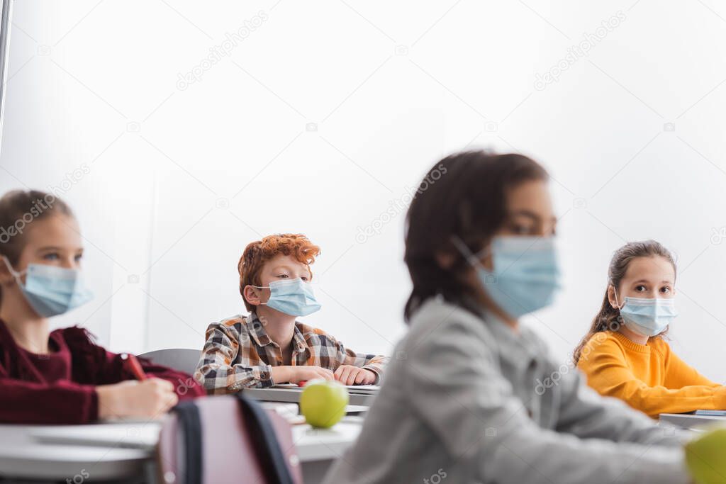 Pupils in medical masks sitting near friends on blurred foreground in classroom 