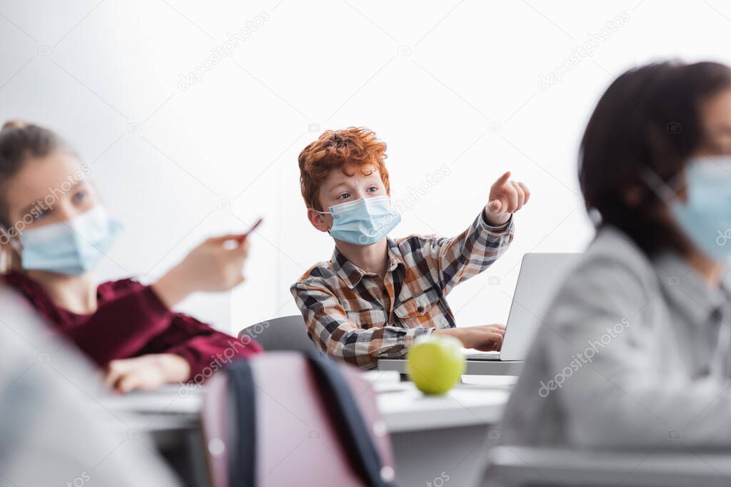 pupils in medical masks pointing with finger and pen during lesson in school, blurred foreground