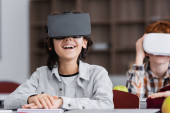 excited boy laughing in vr headset near classmate on blurred background