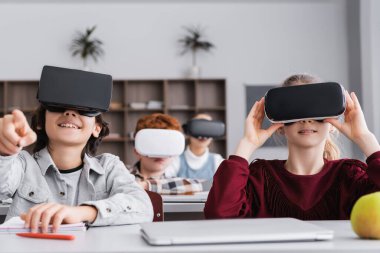 VR kulaklıklı heyecanlı çocuk bulanık arka planda parmaklarıyla sınıf arkadaşlarını işaret ediyor.