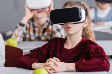 Bulanık arka planda, sınıf arkadaşlarının yanında vr kulaklıkla oynayan liseli kızlar.