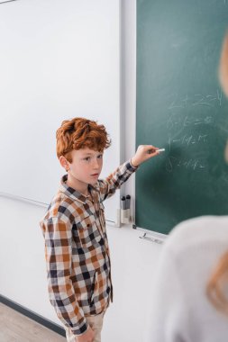 Kızıl saçlı okul çocuğu karatahtaya yazı yazıyor, öğretmeninin yanında, bulanık önplanda.