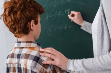 Öğretmen, karatahtaya bulanık arka planda yazı yazarken kızıl öğrencinin omzuna dokunuyor.