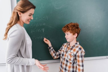 Gülümseyen öğretmen, elleri ile karatahtaya yazı yazan kızıl öğrenciyi işaret ediyor.