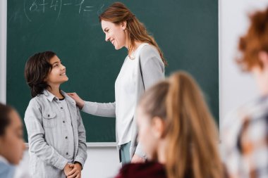 Mutlu öğretmen ve okul çocuğu karatahta ve ön plandaki bulanık öğrencilerin yanında birbirlerine bakıyorlar.