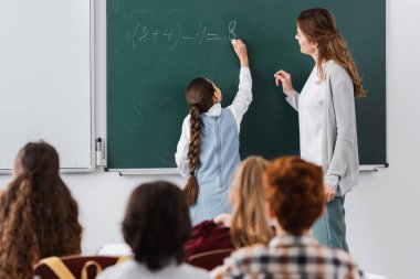 teacher near schoolgirl writing on chalkboard, and pupils on blurred foreground clipart