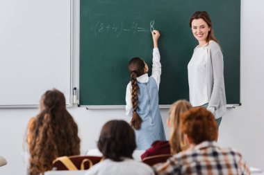 Gülümseyen öğretmen ve bulanık ön planda öğrencilerin olduğu tahtaya yazı yazan okul kızlarının arka planı
