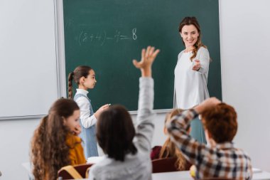 smiling teacher standing and chalkboard and pointing at pupils on blurred foreground clipart