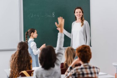 Gülümseyen öğretmen, bulanık ön planda elini kaldıran öğrenciye bakıyor.