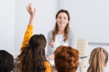 Öğrencilerin yanında elini kaldıran kız öğrenci ve bulanık arka planda öğretmen.