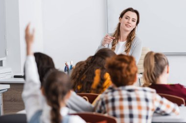 Gülümseyen öğretmen, bulanık ön planda elini kaldıran öğrenciyi işaret ediyor.