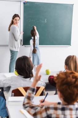 Öğretmen bulanık ön planda elini kaldıran öğrenciye bakıyor.