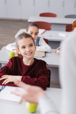 smiling schoolgirl looking at teacher on blurred foreground during lesson clipart