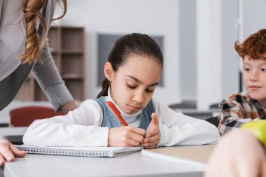 Öğrenci kız ön planda öğretmen ve sınıf arkadaşlarının yanına not defteri yazıyor.