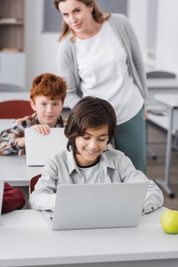 Öğretmen ders sırasında dizüstü bilgisayar kullanarak öğrencilerin yanında duruyor, arkaplan bulanık