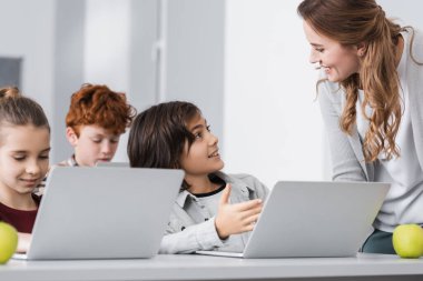 Okullu çocuk sınıfta gülümseyen öğretmenin yanında dizüstü bilgisayarı gösteriyor.