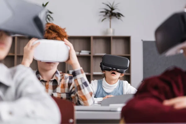 astonished schoolgirl gaming in vr headset near classmates on blurred foreground