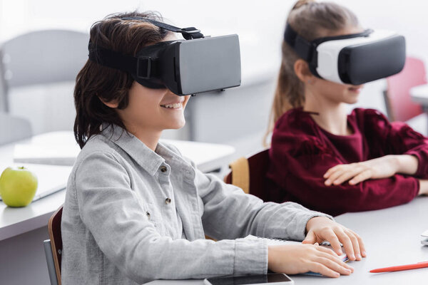 cheerful schoolboy in vr headset near classmate on blurred background