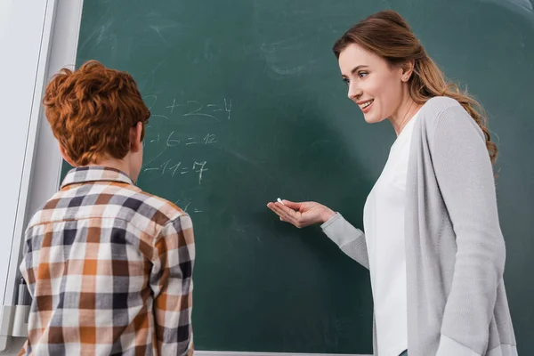 smiling teacher pointing at chalkboard near redhead schoolboy