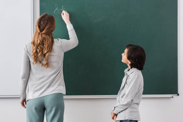 back view of teacher writing on chalkboard near schoolboy