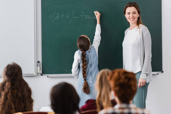 cheerful teacher near schoolgirl writing on chalkboard and pupils on blurred foreground