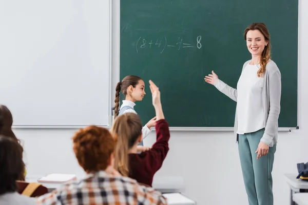 happy teacher pointing at chalkboard near pupils on blurred foreground