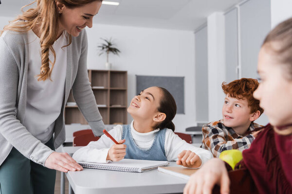 happy schoolgirl looking at smiling teacher in classroom near pupils on blurred foreground 