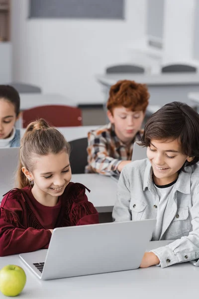 cheerful classmates near laptops, apple and pupils on blurred background