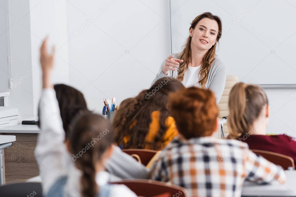 smiling teacher pointing with finger at schoolgirl raising hand on blurred foreground