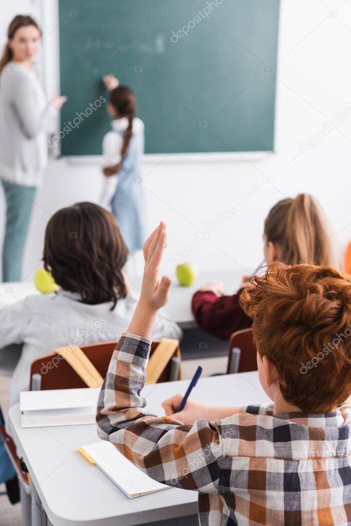 back view of redhead schoolboy raising hand during lesson near teacher on blurred background