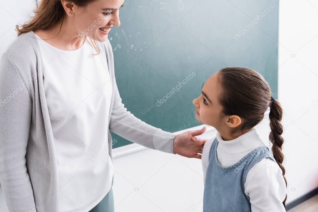 happy schoolgirl and teacher looking at each other near chalkboard