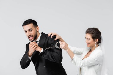 Gülümseyen gelin, gri alanda izole edilmiş korkmuş Müslüman damadın kelepçelerini tutuyor. 