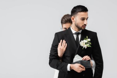 Bride taking dollars from wallet in hand of muslim groom isolated on grey  clipart