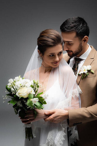 Muslim groom hugging smiling bride with wedding bouquet isolated on grey 