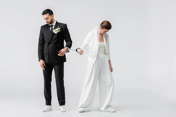 Sad interracial groom and bride in handcuffs on white background