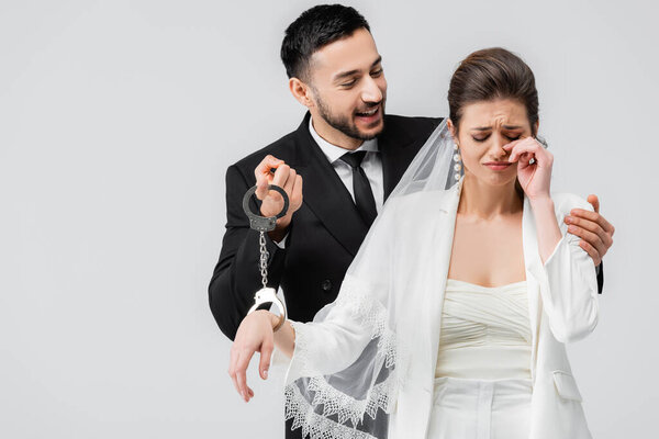 Улыбающийся арабский жених держит наручники рядом с плачущей невестой, изолированной на серой 