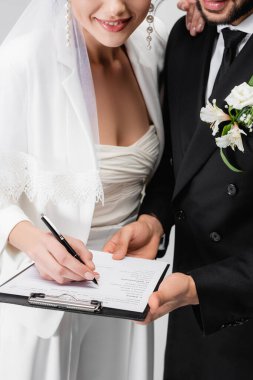 Cropped view of smiling bride signing wedding contract near groom isolated on grey  clipart