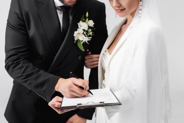 Cropped view of groom signing contract near bride in veil isolated on grey  clipart