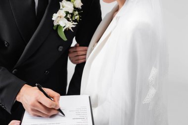 Cropped view of groom signing wedding contract near young bride isolated on grey  clipart