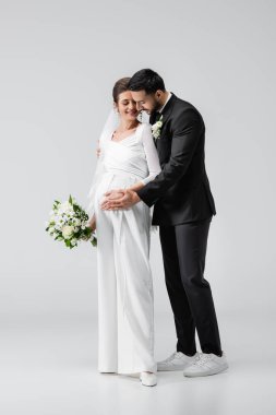 Arap damat, gri arka planda buketle hamile gelini kucaklıyor. 