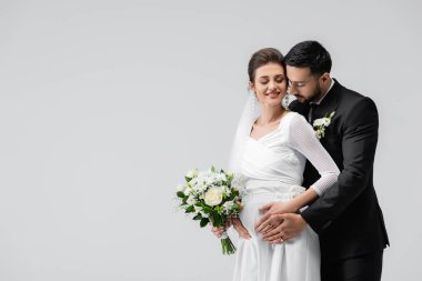 Takım elbiseli Müslüman damat, gülümseyen hamile geline sarılıyor. 