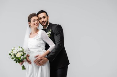Gülümseyen hamile gelin, gri renkte yalnız başına Müslüman damadın yanında duruyor. 
