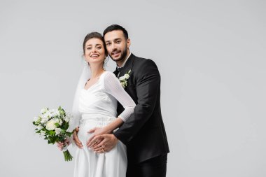 Genç Müslüman damat, gri renkte izole edilmiş düğün buketiyle neşeli hamile gelini kucaklıyor. 
