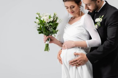 Gri renkte izole edilmiş düğün buketiyle hamile gelinin karnına bakan Müslüman damat. 