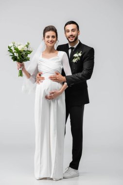 Mutlu Arap damat hamile geline sarılıyor ve gri arka plandaki kameraya bakıyor. 