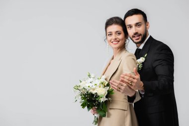 Neşeli çoklu etnik evliler kameraya bakarken gri renkte izole edilmiş alyansları gösteriyorlar.