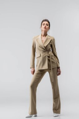full length view of elegant woman in beige pantsuit looking at camera on grey clipart
