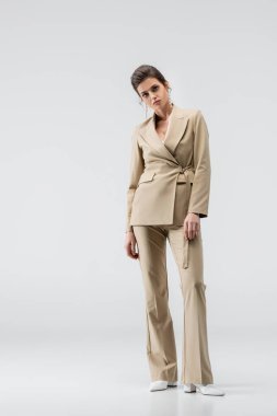 young brunette woman in stylish pantsuit looking at camera while standing on grey clipart