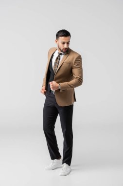 Modaya uygun takım elbiseli, gri renkli spor ayakkabılı esmer Arap adamın tam boy görüntüsü.