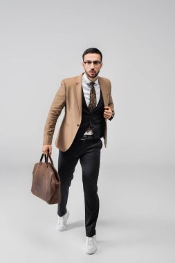 Moda düşkünü Müslüman bir adamın deri çantayla yürüyüp gri renkli kameraya baktığı uzun bir manzara.